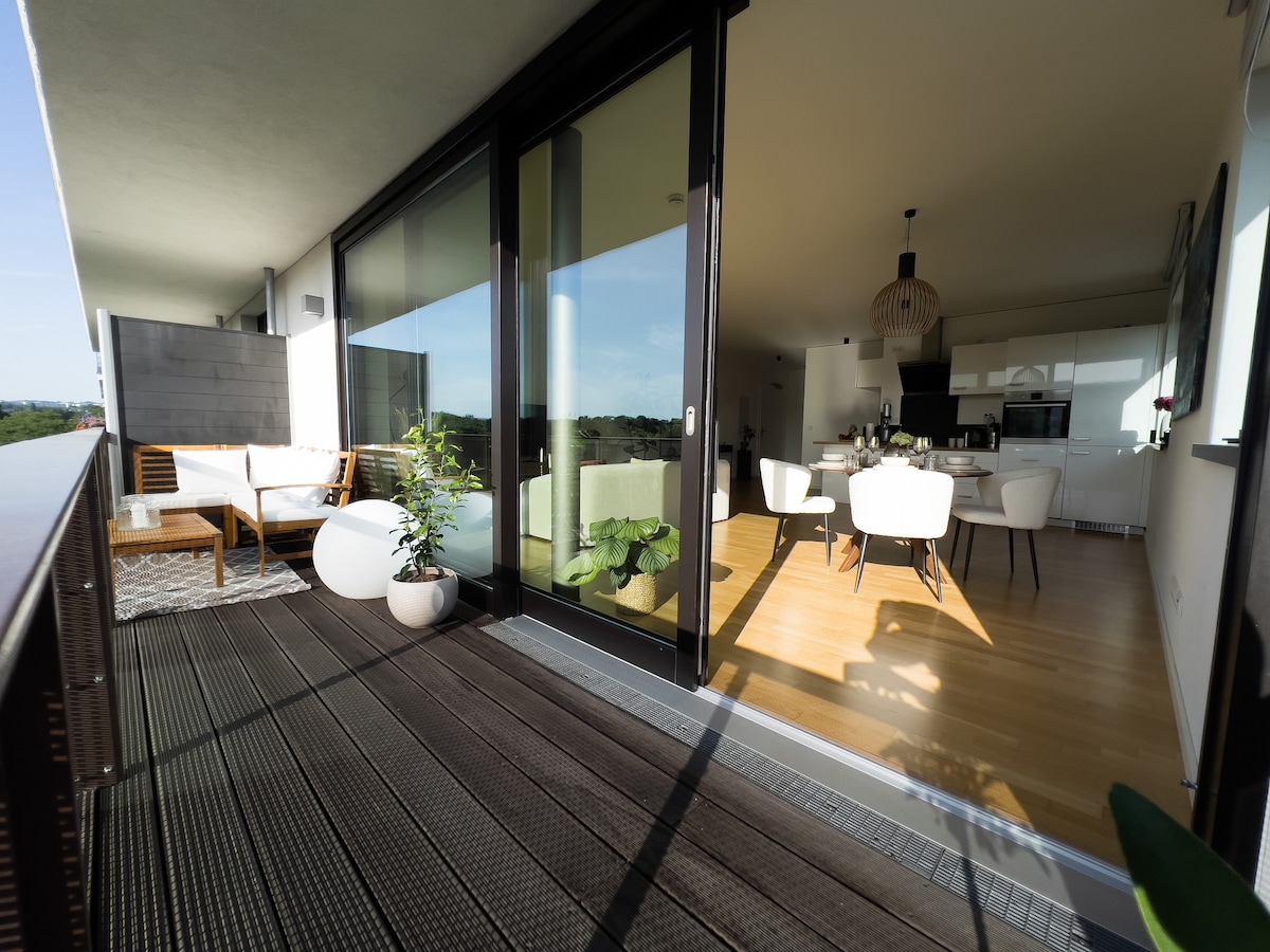 A spacious balcony is visible, featuring outdoor seating with a small table and a decorative plant. Large glass doors lead into a modern, well-lit living area and kitchen. The warm wood floors and open layout create an inviting atmosphere, with views of the surrounding landscape.
