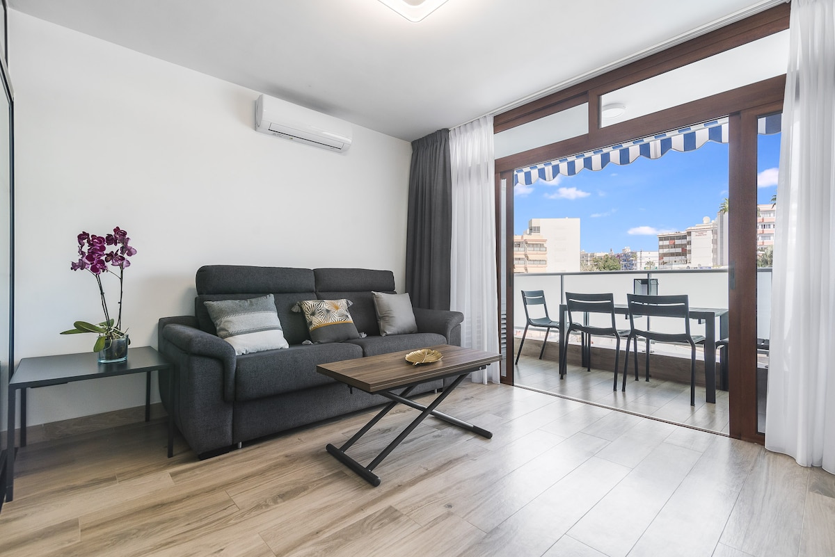 A modern living space is presented with a comfortable gray sofa adorned with decorative pillows. A wooden coffee table is positioned in front of the sofa. Floor-to-ceiling windows showcase an outdoor balcony with a dining area for four, surrounded by city views.