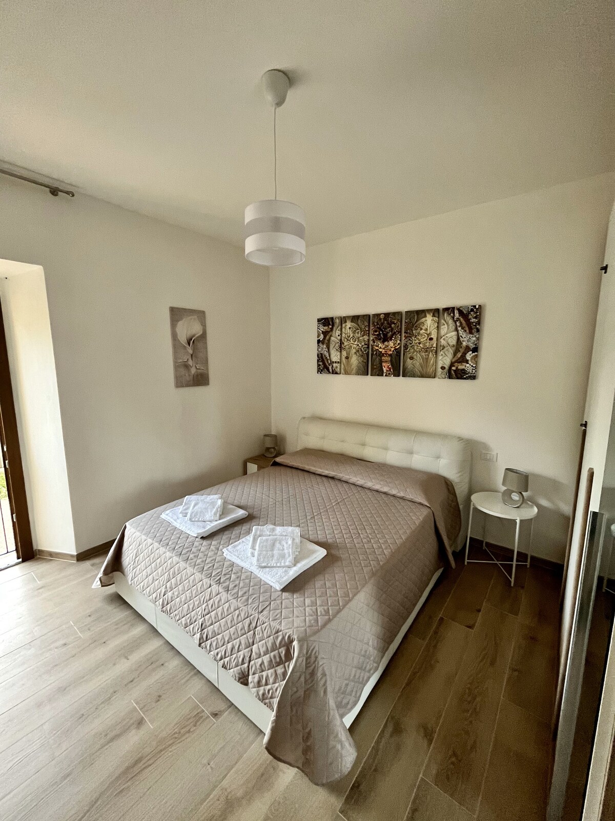 A serene bedroom features a spacious bed covered with a light quilt. Two neatly arranged towels are placed on top. Wall art depicting nature enhances the ambiance, and a bedside table stands beside the bed. Natural light enters through a window, complemented by simple overhead lighting.