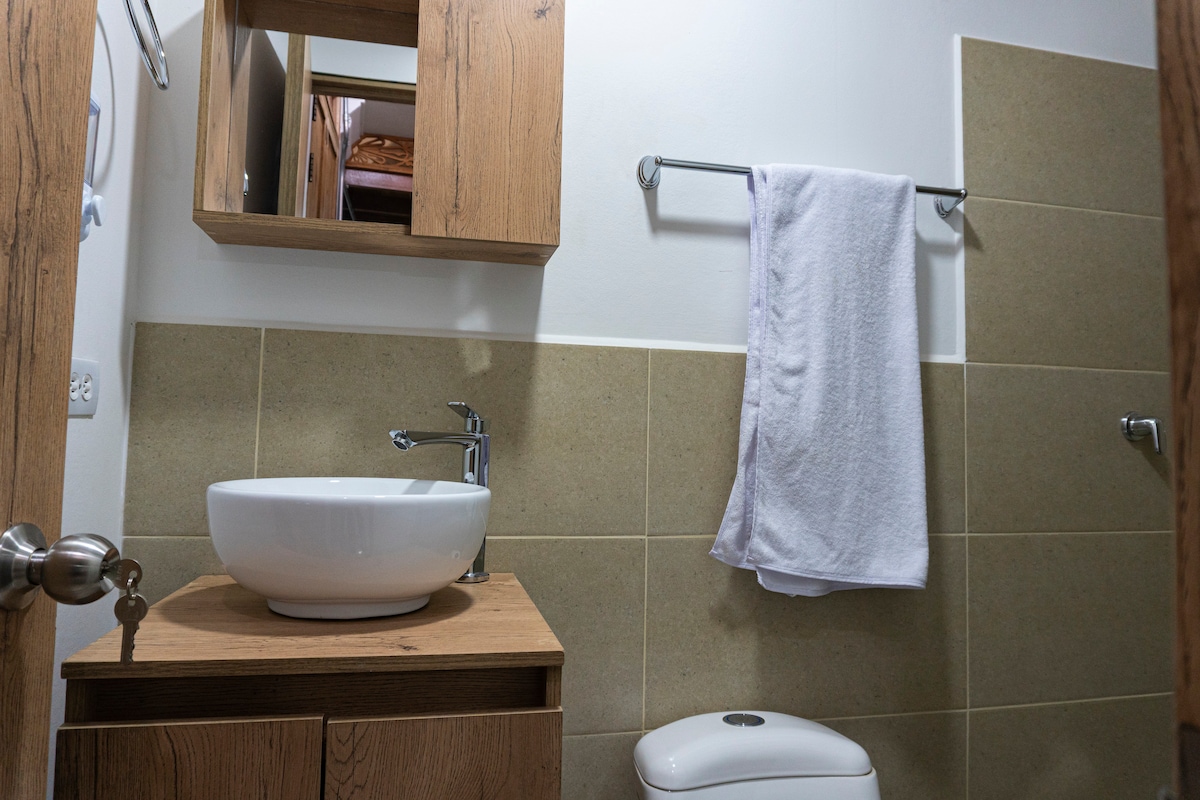 A modern bathroom featuring a wall-mounted wooden cabinet, a round vessel sink on a wooden vanity, and a wall-mounted towel rack. Soft lighting highlights the neutral-toned tiles and a white toilet, creating a clean and functional space.