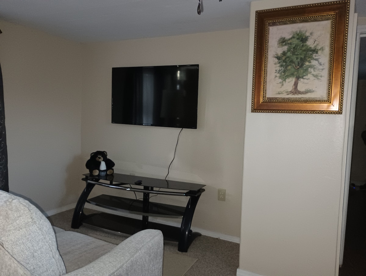 A wall-mounted television is positioned above a sleek black glass stand. A plush couch faces the TV, while a framed artwork featuring a tree hangs on the wall. Soft carpet covers the floor, contributing to a comfortable living space.