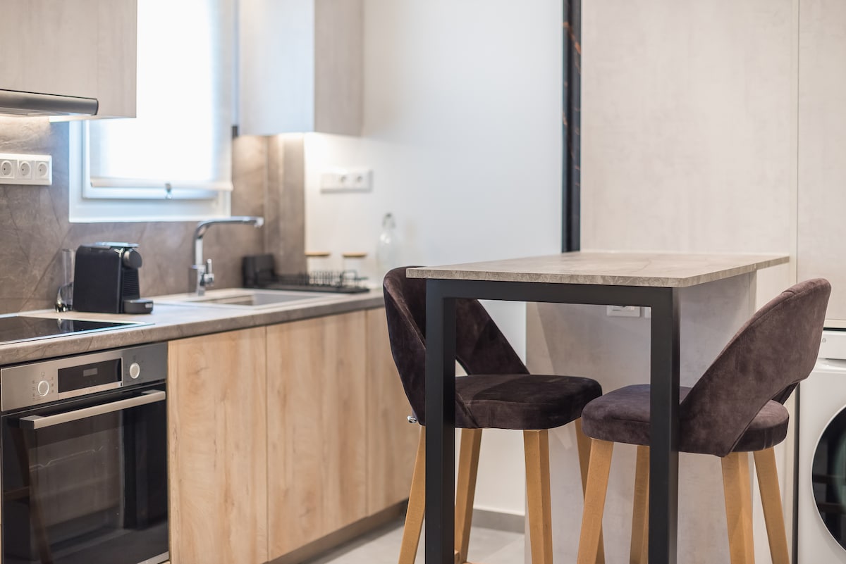 A modern kitchen space is presented, featuring sleek cabinetry in wood and neutral tones. A compact dining table with two upholstered chairs is situated near the window, while essential appliances, including an oven and a washing machine, are integrated into the design.