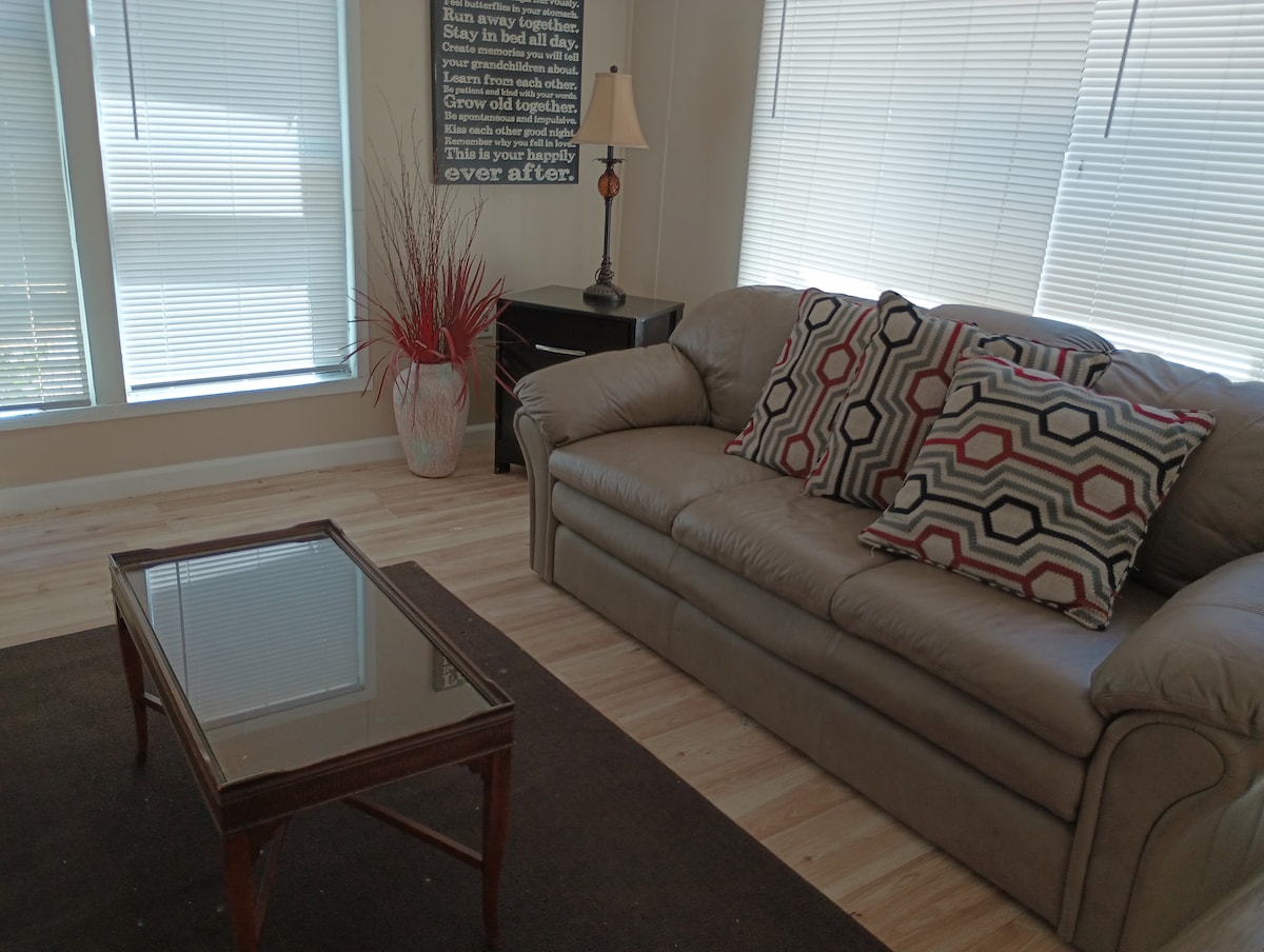 A comfortable living area is showcased with a light-colored sofa adorned with patterned cushions. A glass-topped coffee table is positioned in front of the sofa, complemented by a decorative lamp and potted plant in the background. Natural light filters through window blinds.