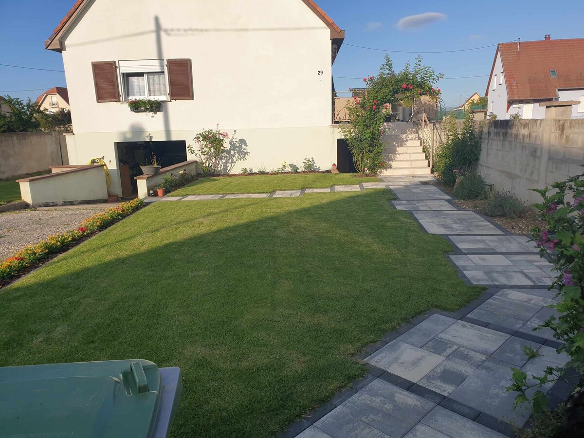 A well-maintained garden area features a neatly trimmed lawn bordered by colorful flower beds. Stone pathways lead towards the entrance of the house, which has large windows with wooden shutters. A flight of stairs is visible, providing access to the upper level.