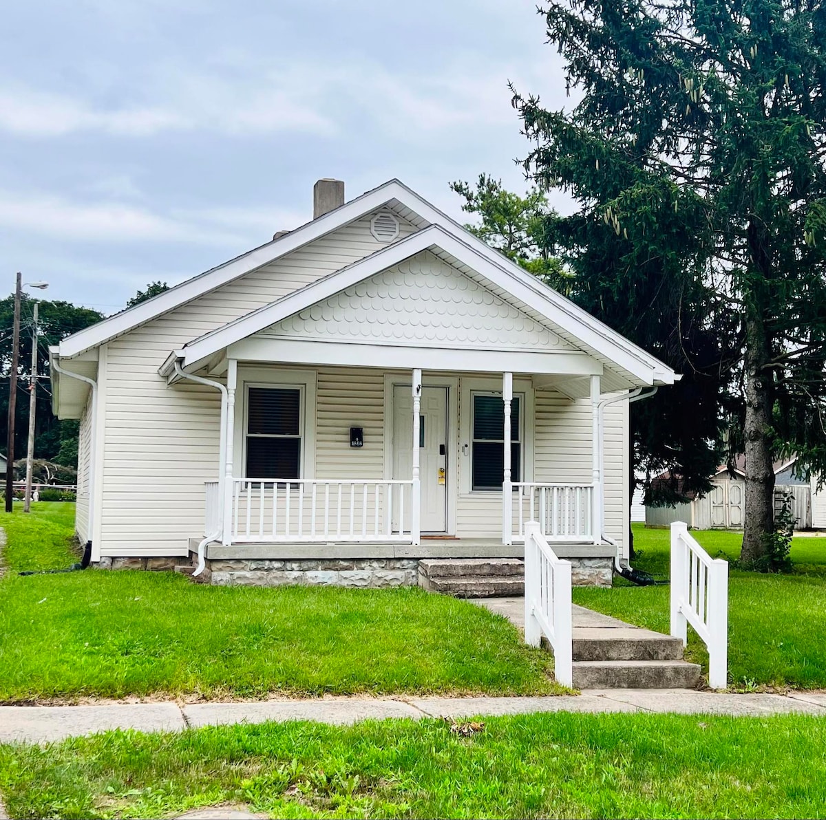 A charming single-story home is presented, featuring a light-colored exterior with distinctive siding. A front porch with a railing enhances accessibility, while a small set of steps leads up to the entrance. Lush green grass surrounds the property, contributing to a welcoming environment.