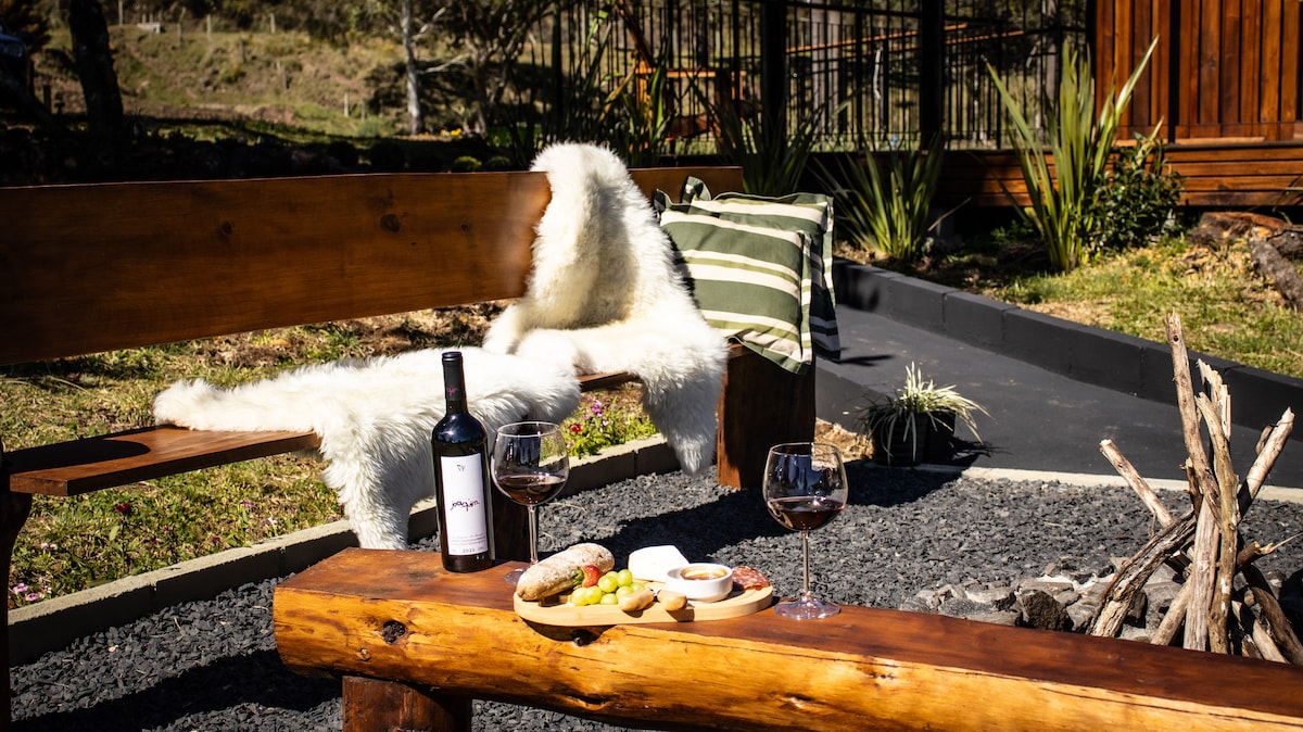 An inviting outdoor seating area features a rustic wooden bench topped with a soft white throw. A wooden table holds two glasses of red wine and a platter of snacks. The background includes lush greenery and a stone path leading to the property.