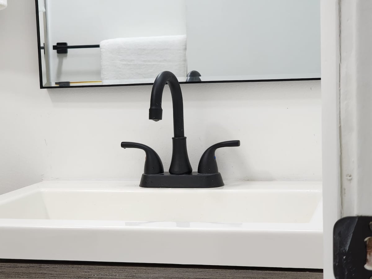 A modern bathroom sink features a sleek black faucet set against a light-colored wall. A large mirror reflects the setup, with neatly arranged towels visible in the background. The sink design is minimalistic, contributing to a clean and tidy space.