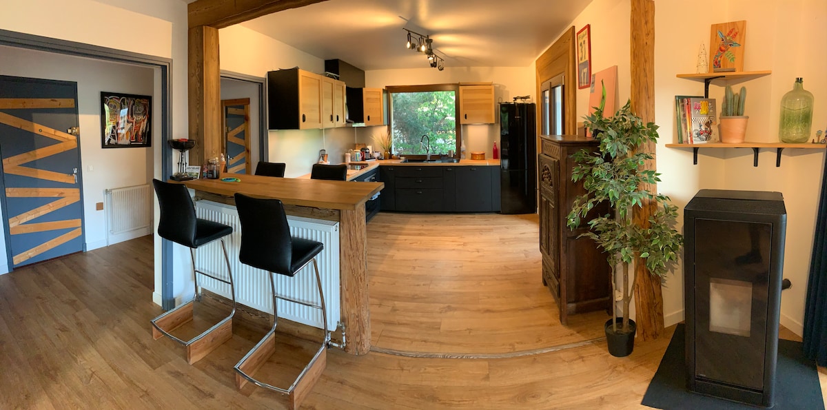 A kitchen area features wooden cabinetry and modern appliances, creating a functional space for meal preparation. A central island with seating for two is present, along with a cozy wood stove on one side. Warm wooden floors contribute to the overall welcoming atmosphere.