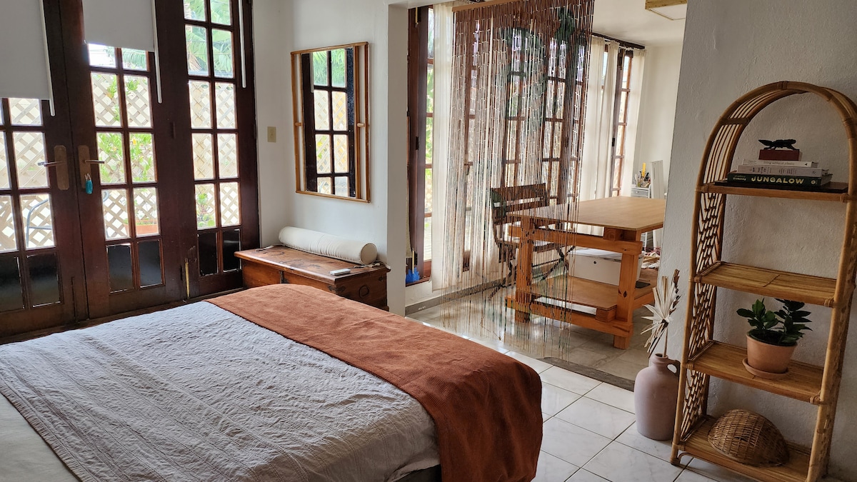 A light-filled space features a spacious bed with a soft, neutral blanket. Natural wood furnishings include a dresser and a dining table set for creative use. Large windows and a beaded curtain enhance the open atmosphere, while potted plants add a touch of greenery.