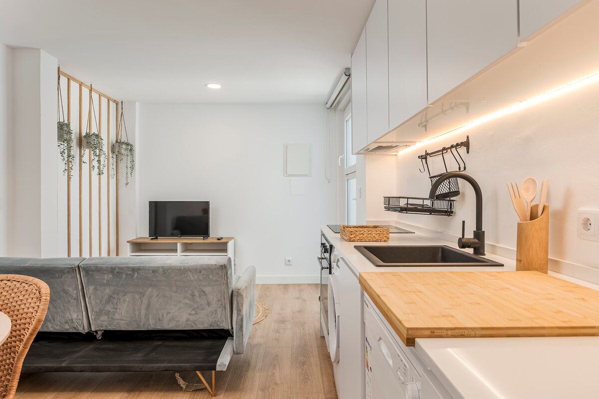 A modern studio space showcases a well-equipped kitchen adjacent to a comfortable living area. A sleek sink and wooden countertop are visible, with cooking utensils neatly arranged. A television sits on a minimalist stand, while a light-filled atmosphere is created by the large windows.