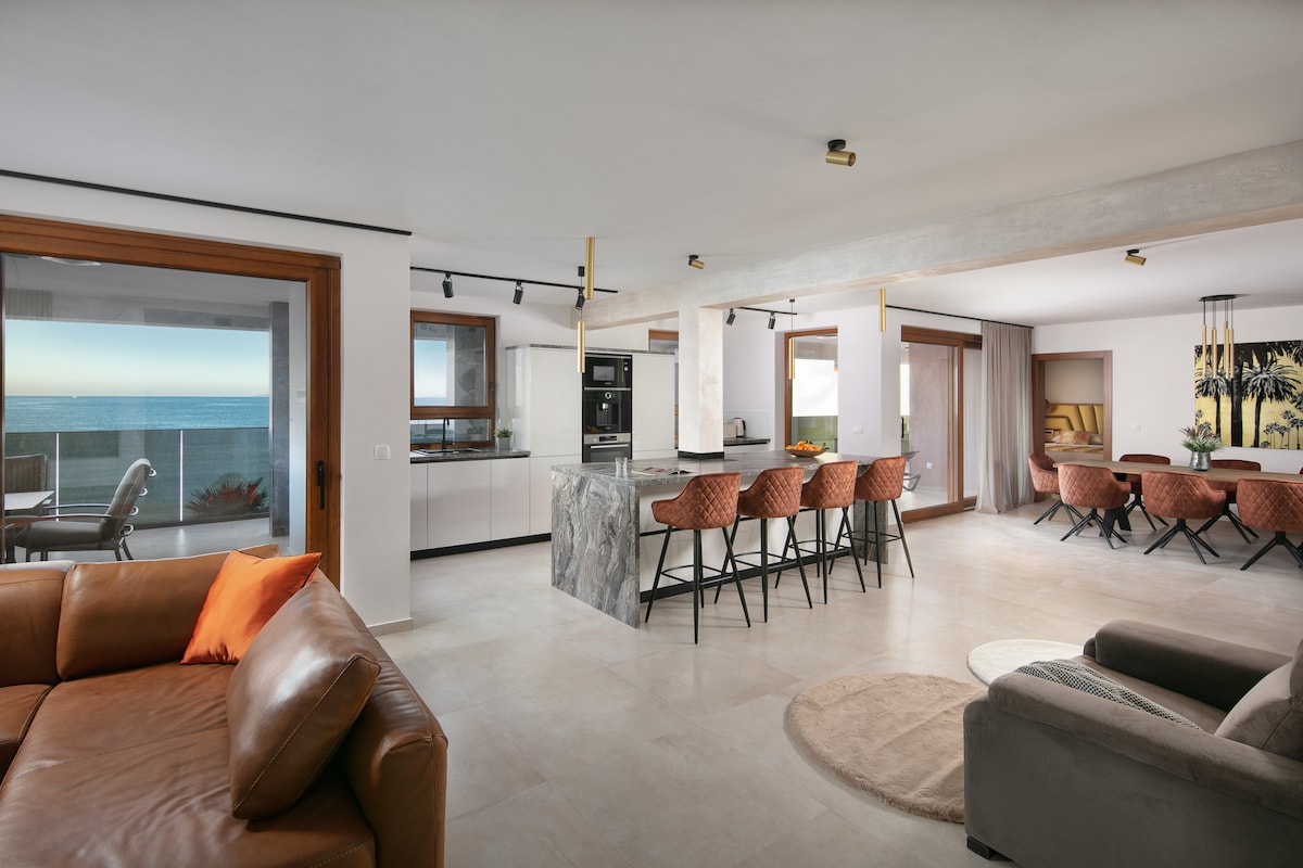 An open-concept living area features sleek marble countertops and modern bar seating. Large windows expose scenic sea views, while a cozy sofa arrangement invites relaxation. A stylish dining space is visible in the background, enhancing the apartment's spacious atmosphere.