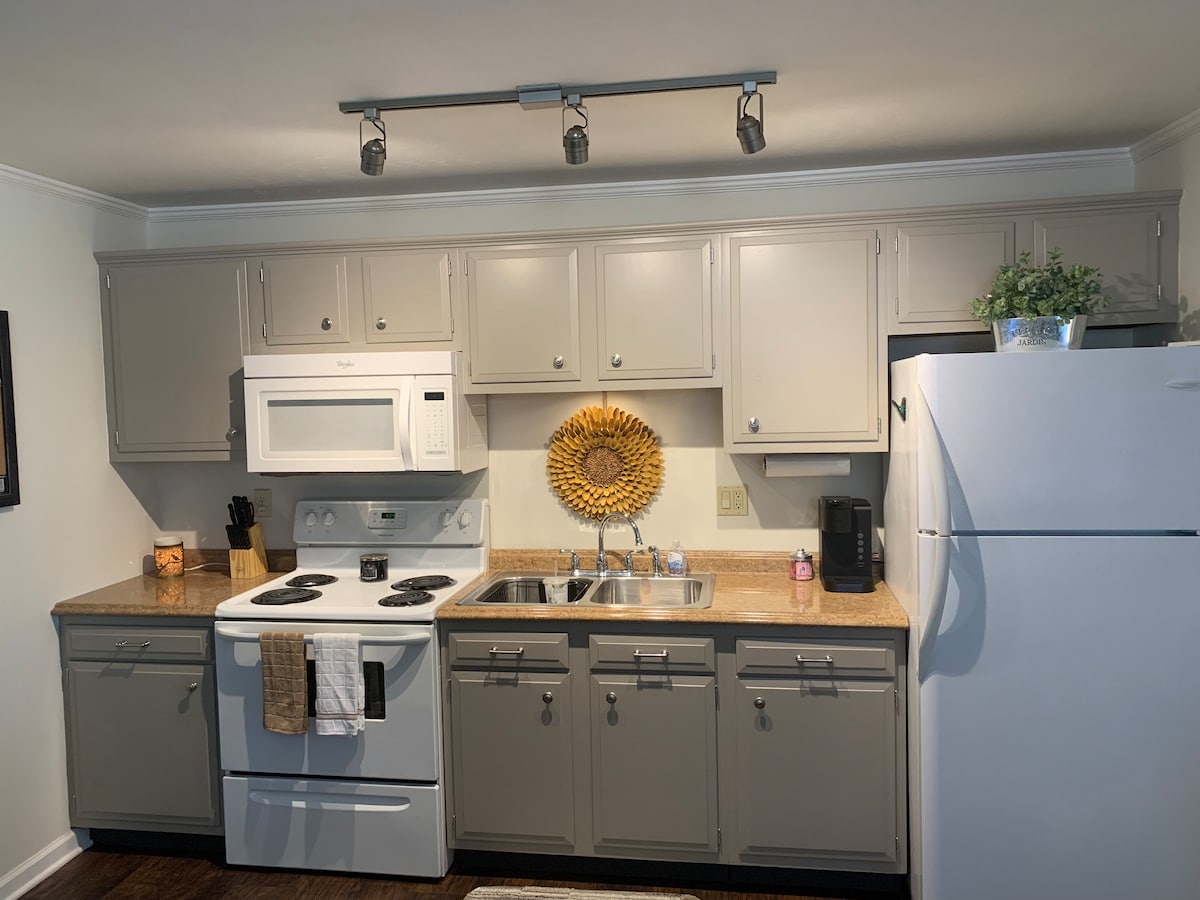 A functional kitchen is equipped with essential appliances, including a stove, microwave, and refrigerator. Light-colored cabinetry complements the granite countertops. A decorative wall piece adds a touch of style above the sink. Natural light illuminates the space through soft-colored walls.