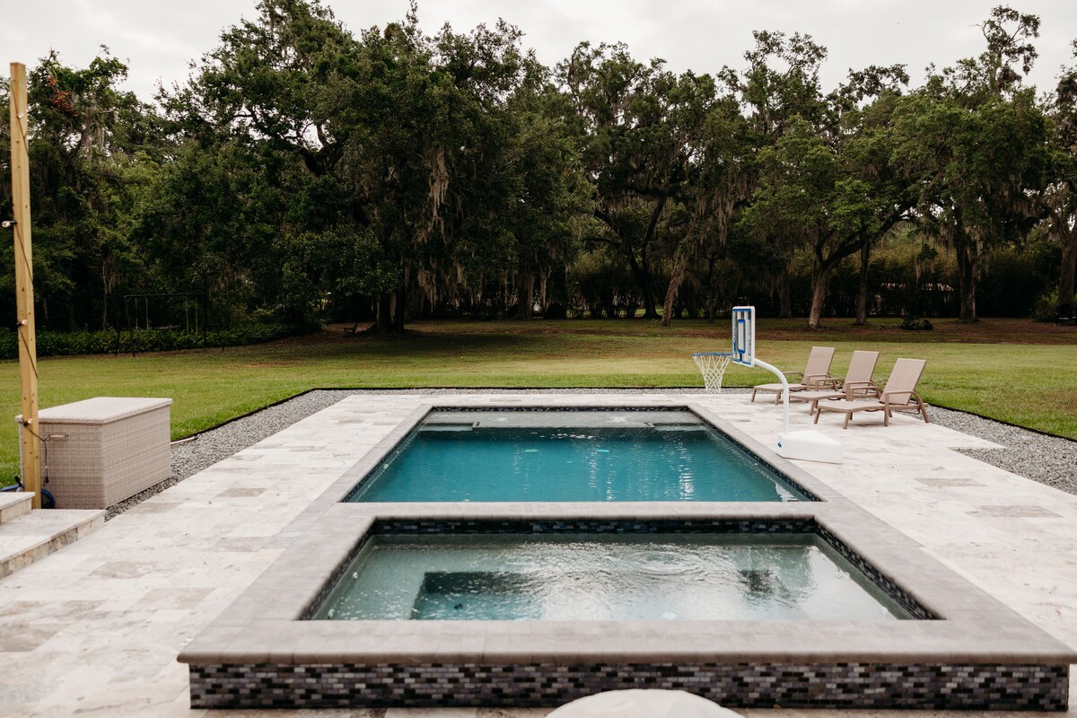 A serene outdoor area features a sparkling swimming pool with a spa on one side, surrounded by a stone patio. Three lounge chairs are positioned nearby, facing a tranquil green landscape with trees in the background.