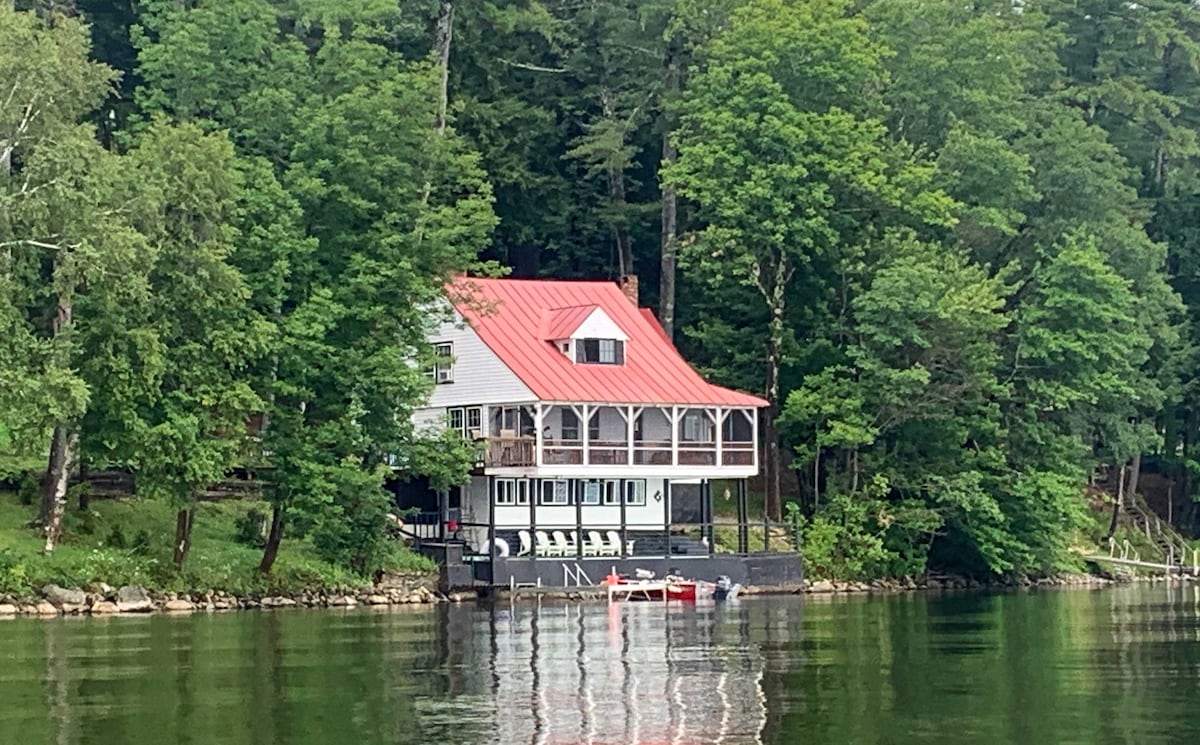 A charming lakefront home is nestled among lush greenery and trees. The structure features a red metal roof and a large screened-in porch that offers expansive seating. The peaceful water reflects the house, enhancing the tranquil ambiance of this scenic location.