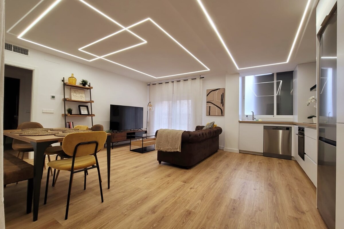The living area features a spacious brown sofa, a dining table with four chairs, and a large television. Natural light filters through sheer curtains, with modern lighting fixtures illuminating the ceiling. The adjacent kitchen area is equipped with stainless steel appliances and sleek cabinetry.