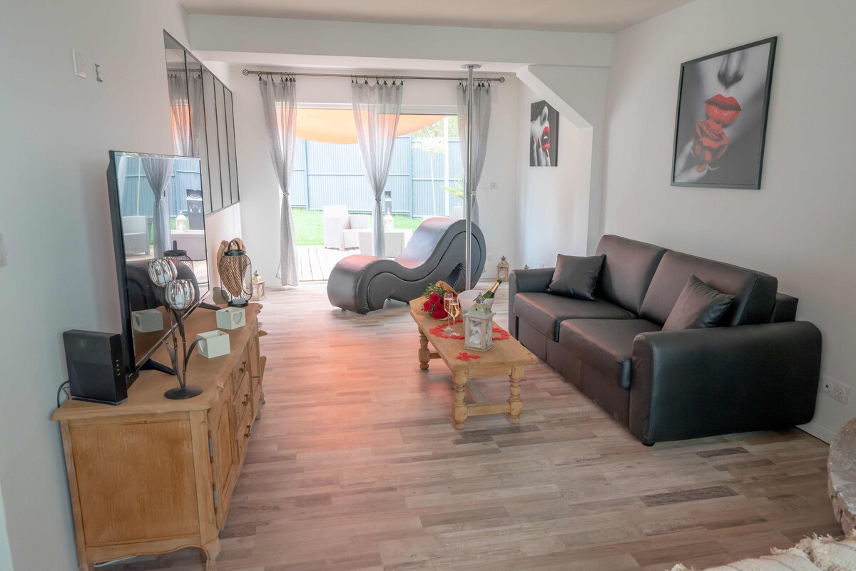 The living area features a sleek black sofa positioned across from a wooden coffee table adorned with floral arrangements. A Tantra sofa is visible, and large windows allow natural light to brighten the space. A wall art piece adds a subtle pop of color.