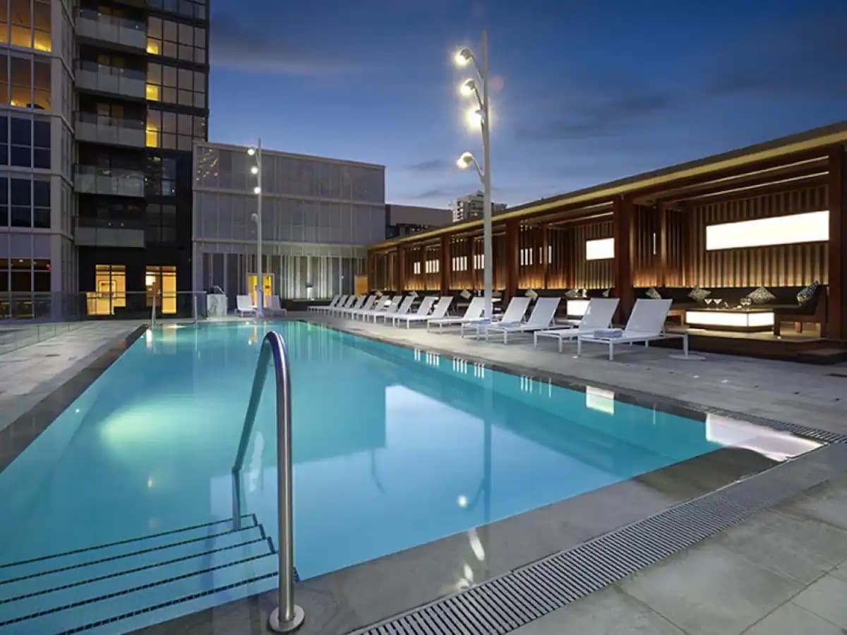An outdoor pool area is illuminated under a twilight sky, featuring a sparkling blue pool surrounded by loungers. Modern seating areas with wooden accents provide a welcoming space for relaxation, while architectural lighting highlights the clean lines of the surrounding buildings.