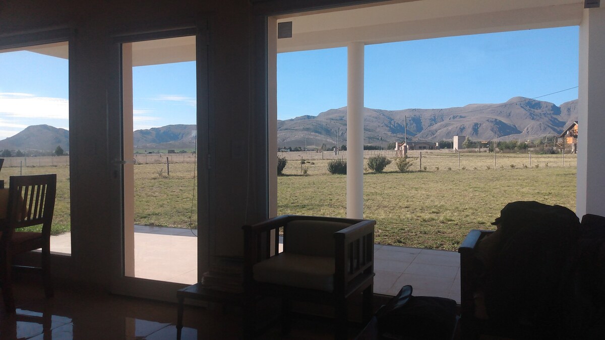 Large windows frame expansive views of the surrounding mountainous landscape, allowing natural light to fill the interior space. A comfortable seating area is visible, featuring wooden furniture that complements the serene environment.