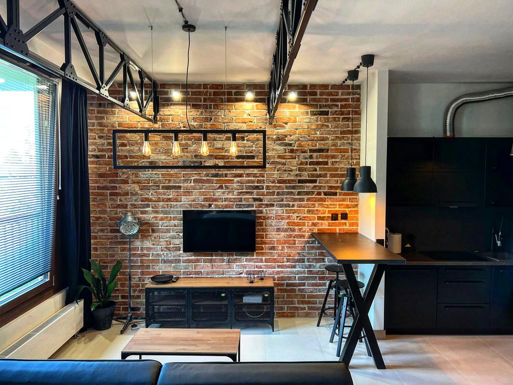An industrial-style living area is highlighted by exposed brick walls and modern pendant lighting. A flat-screen TV is mounted on the wall, while a sleek kitchen area with dark cabinetry is visible in the background. A potted plant adds a touch of greenery.
