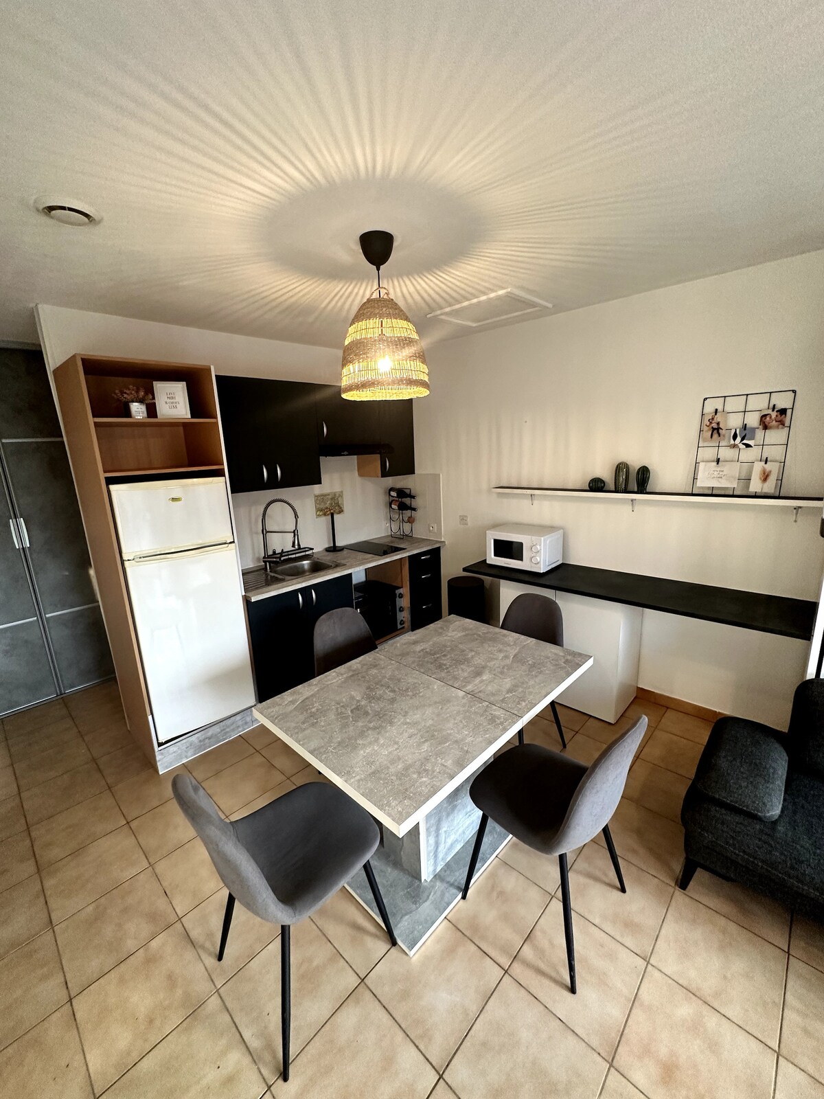 A modern studio kitchen is shown, featuring sleek black and light wood cabinetry. A circular dining table is surrounded by four upholstered chairs. A microwave and refrigerator are positioned on the countertop, with soft lighting creating a welcoming ambiance.