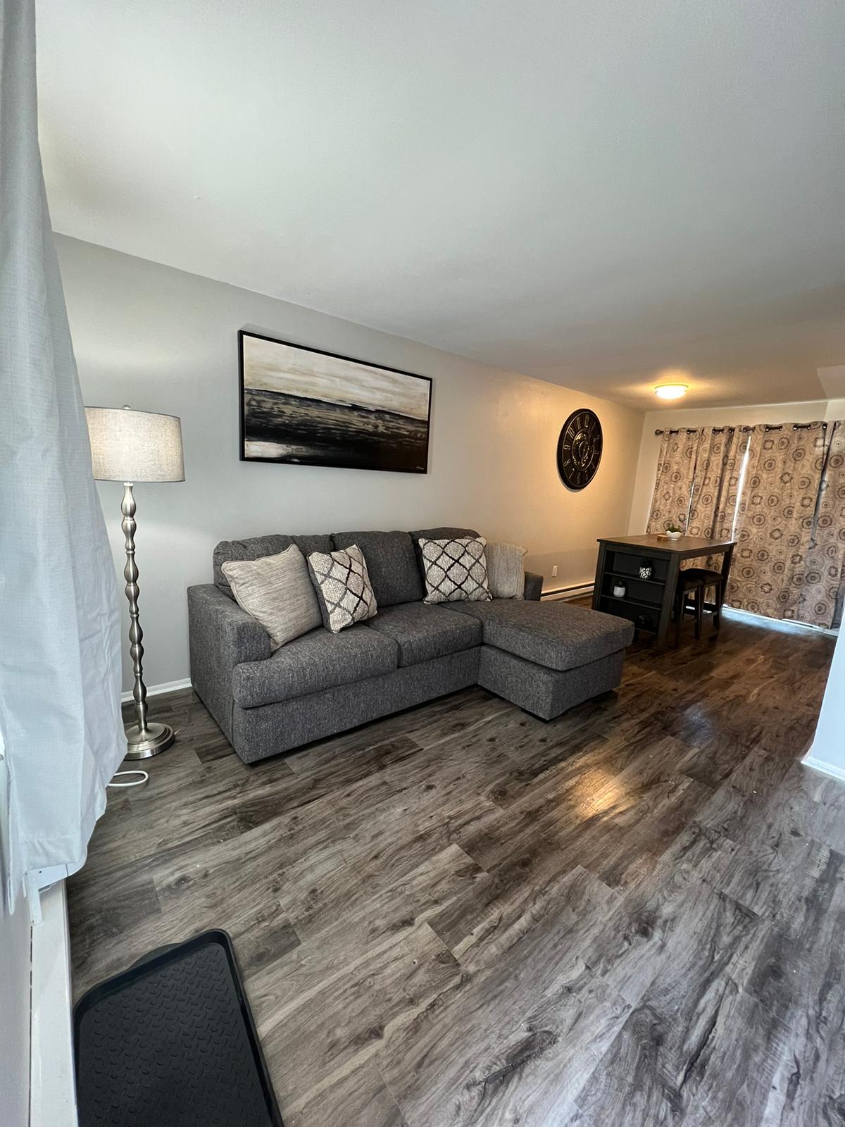 A living area features a gray sectional sofa adorned with decorative pillows, positioned next to a floor lamp. A small dining table is visible, with patterned drapes filtering natural light from the window. Warm wood-like flooring extends throughout the space.
