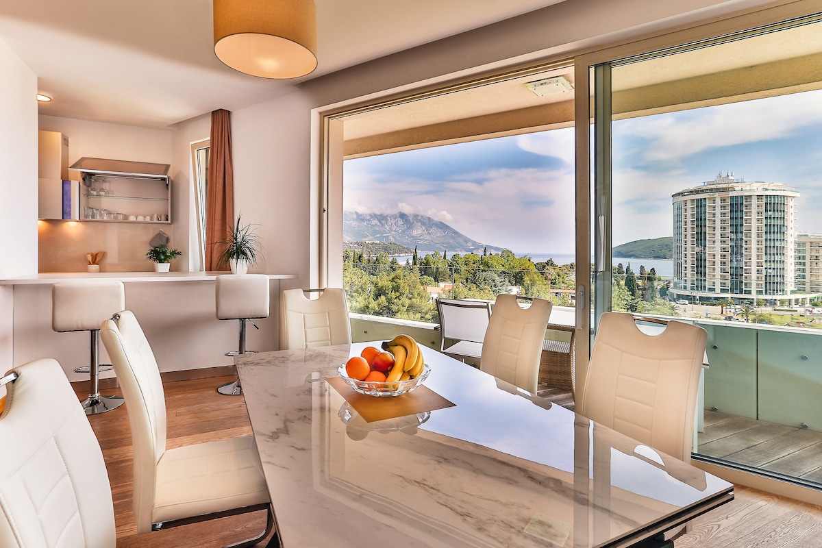 A dining area is featured with a large marble table surrounded by white chairs. An assortment of fresh fruit is displayed in a glass bowl at the center. Expansive windows offer views of the outdoors, enhancing the light-filled atmosphere of the space.