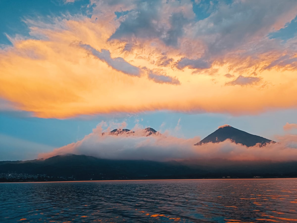 The image captures a stunning view of Lake Atitlan, with two majestic volcanoes rising in the background. Vibrant clouds painted in hues of orange and blue reflect against the water's surface, creating a serene atmosphere that highlights the beauty of the surrounding landscape.