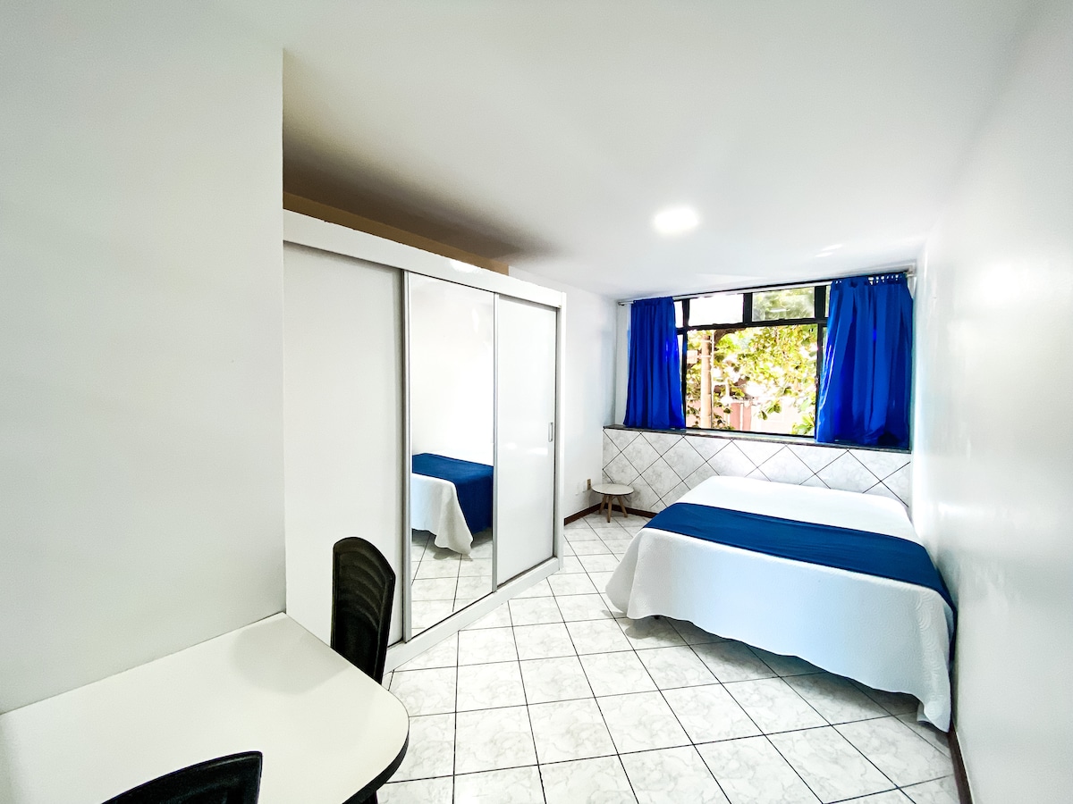 A bright room features a spacious bed with a white cover, complemented by blue curtains. A mirrored wardrobe enhances the room's openness, while a small desk and chair provide a functional workspace. Natural light filters in through a large window, creating an airy ambiance.