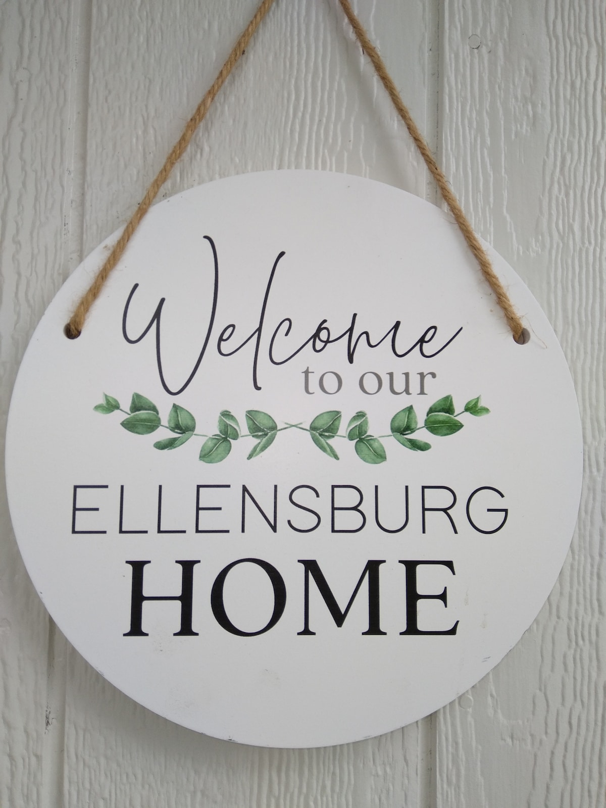 A circular wooden sign displays the message 'Welcome to our Ellensburg HOME,' featuring elegant black lettering on a white background. Green leafy accents are incorporated into the design, adding a touch of nature to the welcoming message.