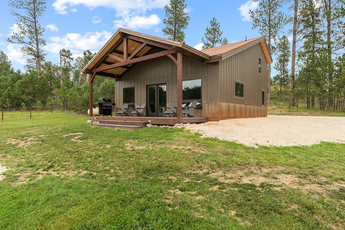 A modern timber frame home is set in a serene natural landscape, surrounded by pine trees. The large patio features comfortable seating, and a gas grill is visible. The home's expansive windows reflect the beautiful outdoor scenery.
