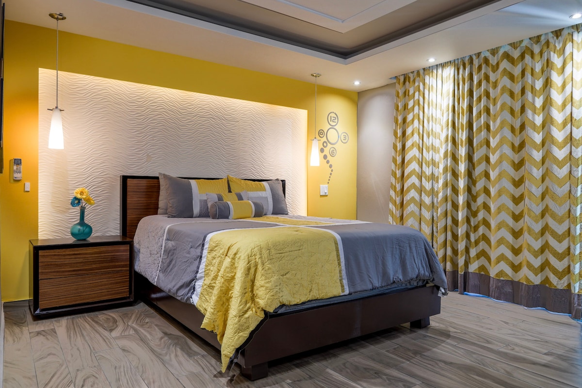 A comfortably arranged bedroom features a spacious bed with yellow and gray bedding. One bedside table holds a vase with flowers. Light from wall sconces and natural light floods the room, complemented by patterned curtains and a textured accent wall.