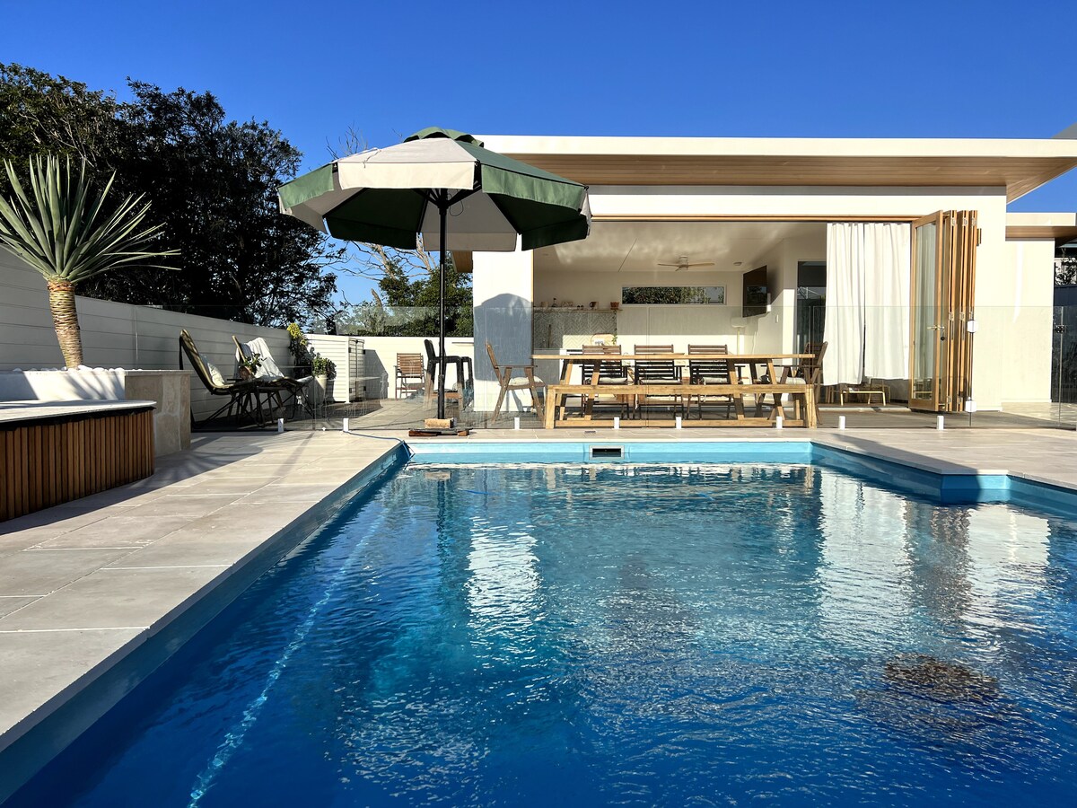 A private pool is surrounded by modern outdoor furniture, including a large dining table beneath a shade umbrella. The clear blue water reflects the sky, while a relaxed seating area is visible in the background, enhancing the inviting outdoor space.