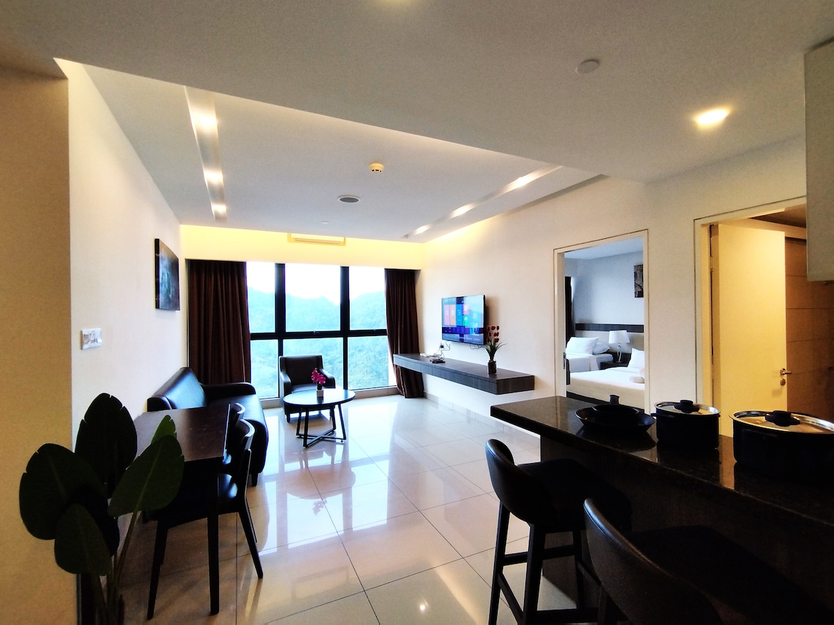 A spacious living area is highlighted by large windows, providing views of surrounding mountains. Modern furnishings include a long leather seating arrangement and a dining table. A 50-inch TV is mounted on the wall, and soft lighting enhances the ambiance.