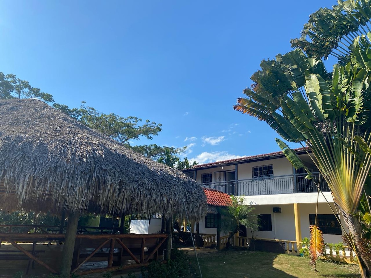 A traditional thatched-roof hut stands in the foreground, surrounded by lush greenery. In the background, a two-story building showcases a terracotta-tiled roof and multiple balconies. The clear blue sky provides a serene backdrop to this inviting outdoor space.