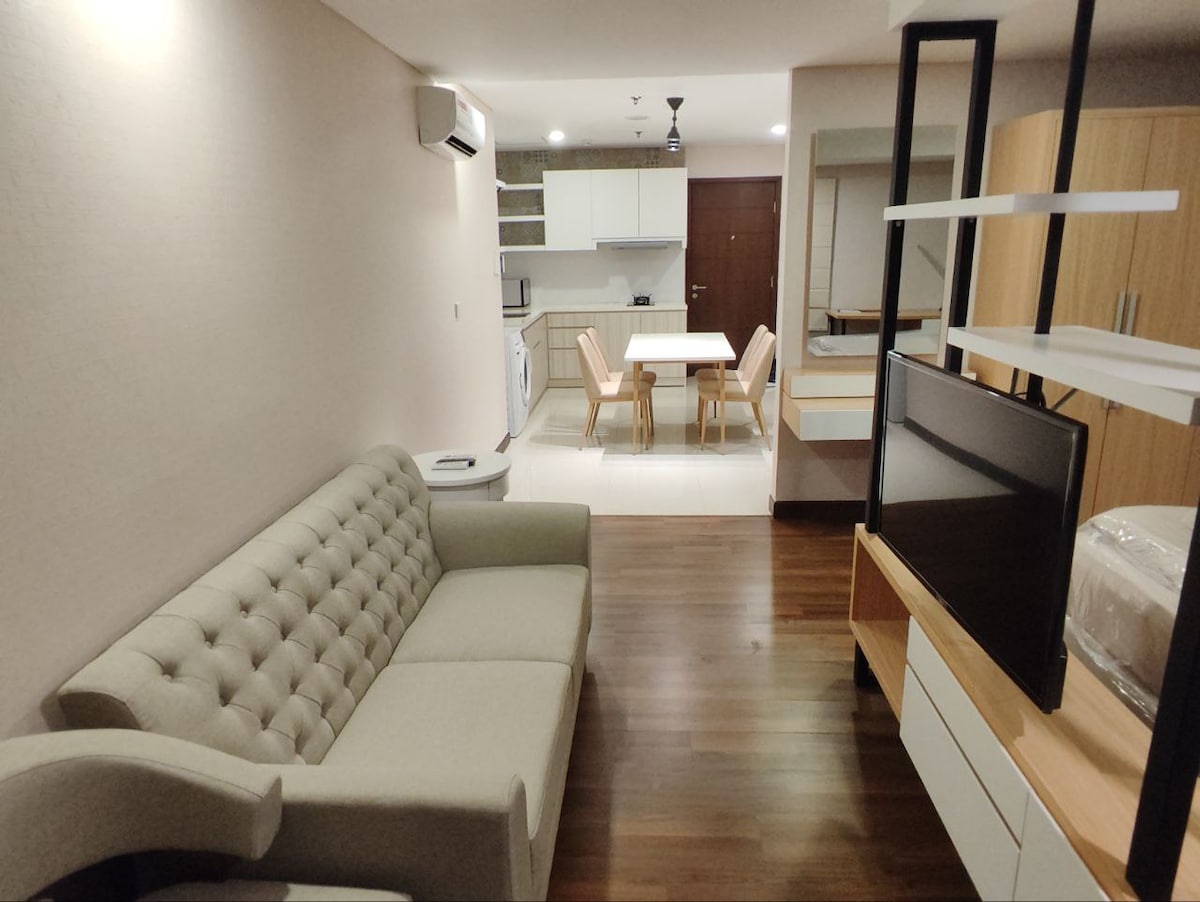 A modern living area features a tufted sofa facing a sleek entertainment unit. A dining table with chairs is positioned in the background, adjacent to a well-equipped kitchen. Warm wooden flooring complements the neutral wall tones, creating a cozy and functional space.