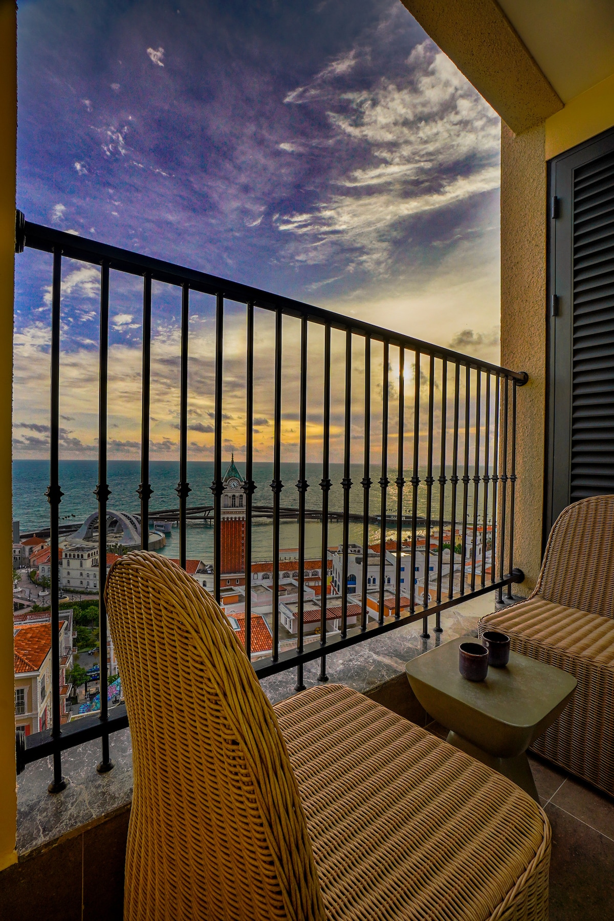 A balcony view is showcased with a railing overlooking the sea and cityscape. Two stylish wicker chairs are positioned around a small table, accompanied by two cups. A serene sky blends hues of orange and blue as the sun sets, creating a tranquil atmosphere.