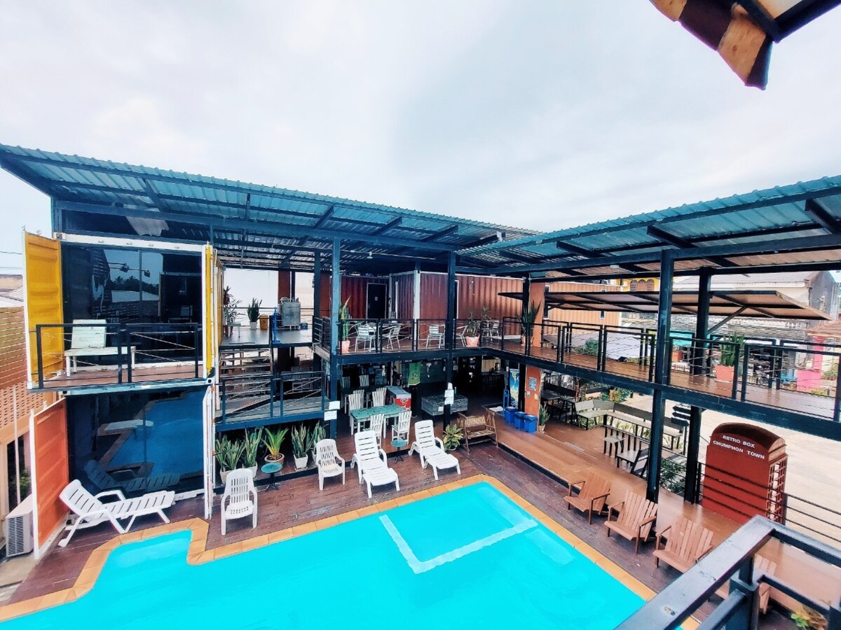 An outdoor swimming pool is surrounded by a wooden deck with lounge chairs and tables. A two-story structure showcases colorful shipping containers and ample covered seating areas. Lush greenery is present throughout the space, enhancing the open, inviting setting.