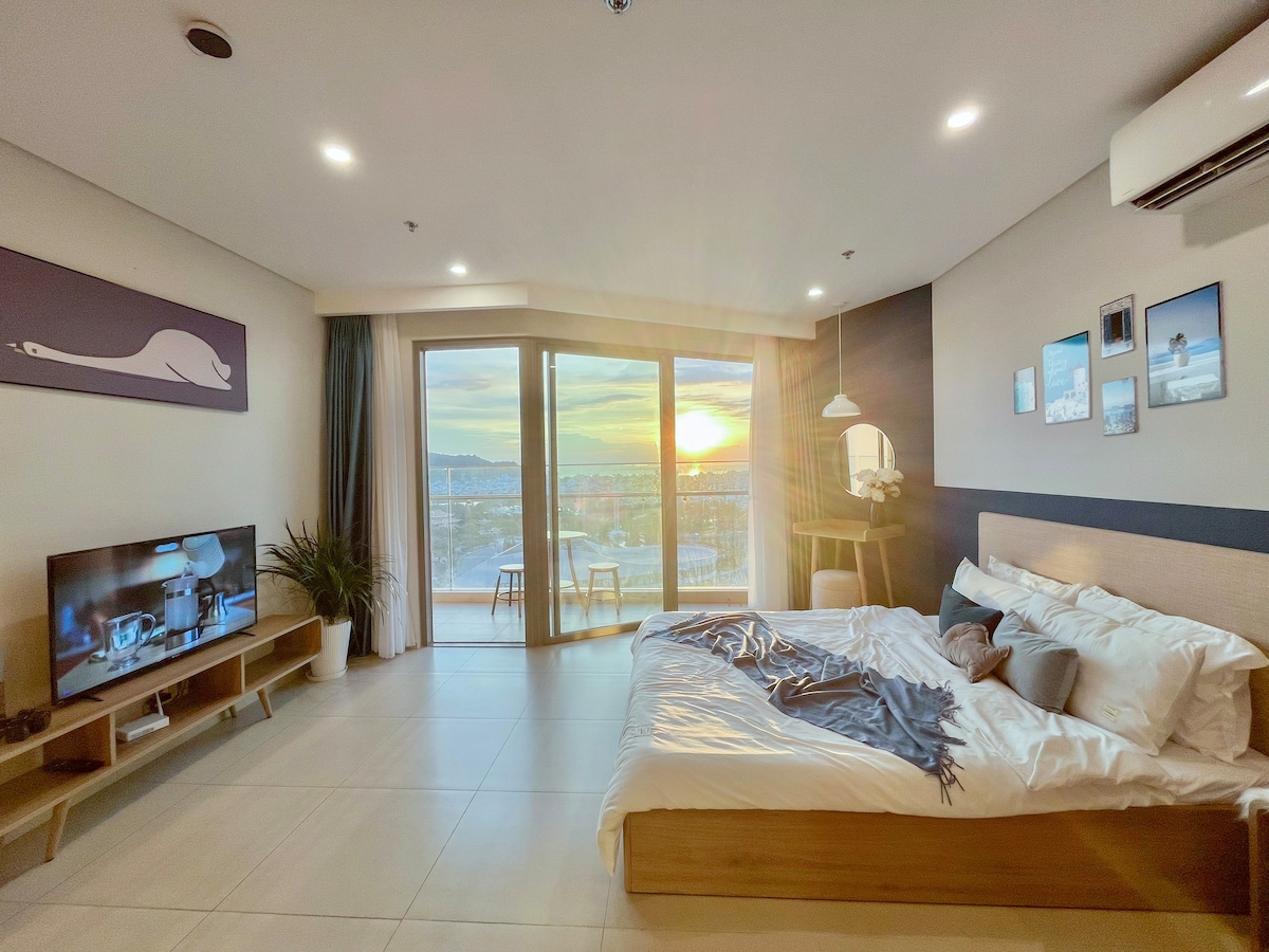 The spacious room features a comfortable bed with crisp white linens, complemented by decorative pillows. A large flat-screen TV is positioned on a wooden console. Large glass doors offer panoramic views of the sunset, while a small dining table and chairs sit on the balcony.