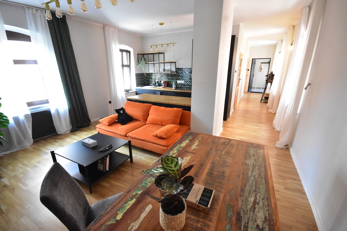 A spacious interior showcases an orange sectional sofa paired with a contemporary black coffee table. A wooden dining table is adorned with a small plant. The kitchen, featuring modern cabinetry, is partially visible in the background, with light streaming through large windows draped in soft curtains.