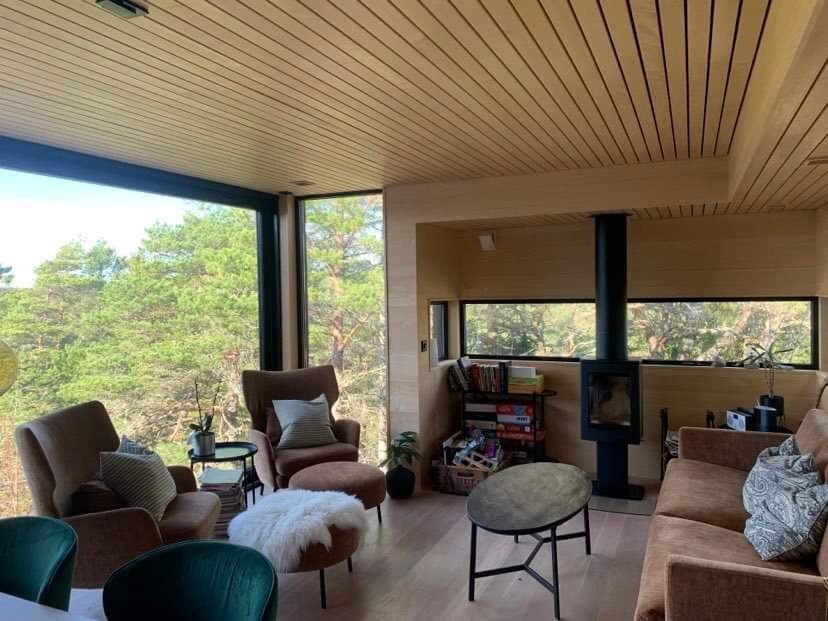 An inviting living area features large windows that frame views of the surrounding trees. Two comfortable armchairs and a green chair are positioned near a small round table. A modern wood stove is set against the wall, and bookshelves display various items.