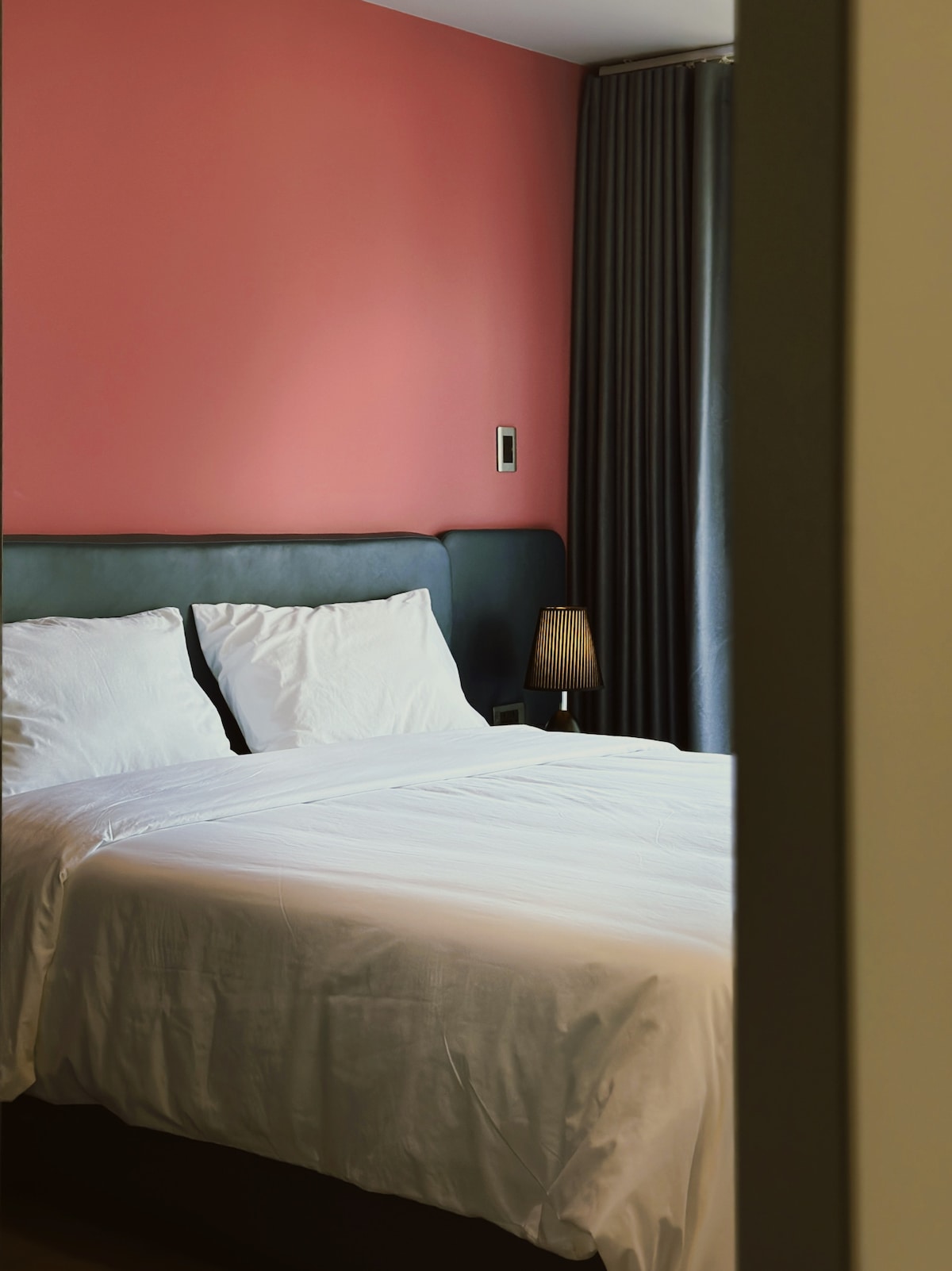 A tranquil bedroom features a spacious bed with neatly arranged white linens and soft pillows. The backdrop consists of a coral-colored wall complemented by dark curtains. A small lamp adds subtle lighting, creating a relaxing atmosphere within the room.