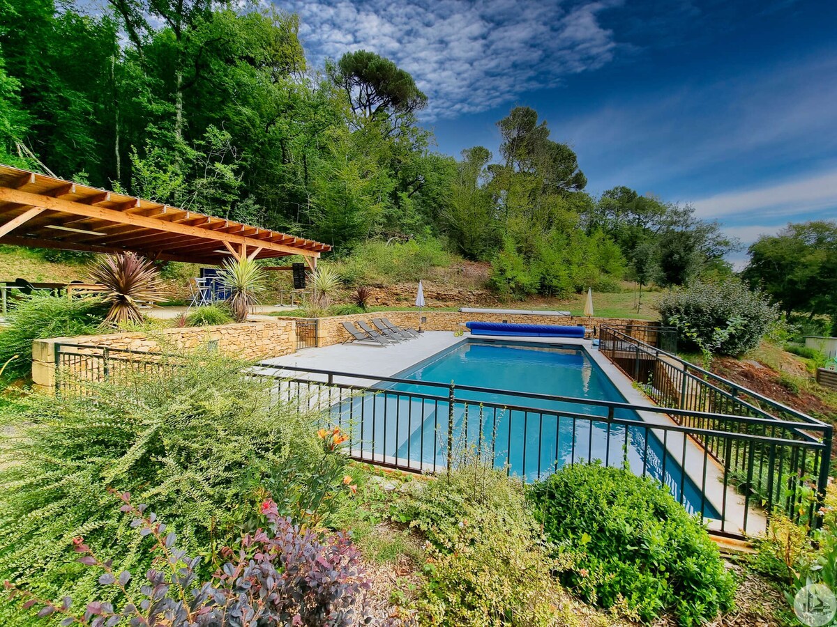 A heated swimming pool, measuring 10x4 meters, is surrounded by stone walls and a metal railing. Lush greenery and various plants enhance the natural setting. A covered area with wooden beams offers shade, complementing the tranquil atmosphere of the outdoor space.