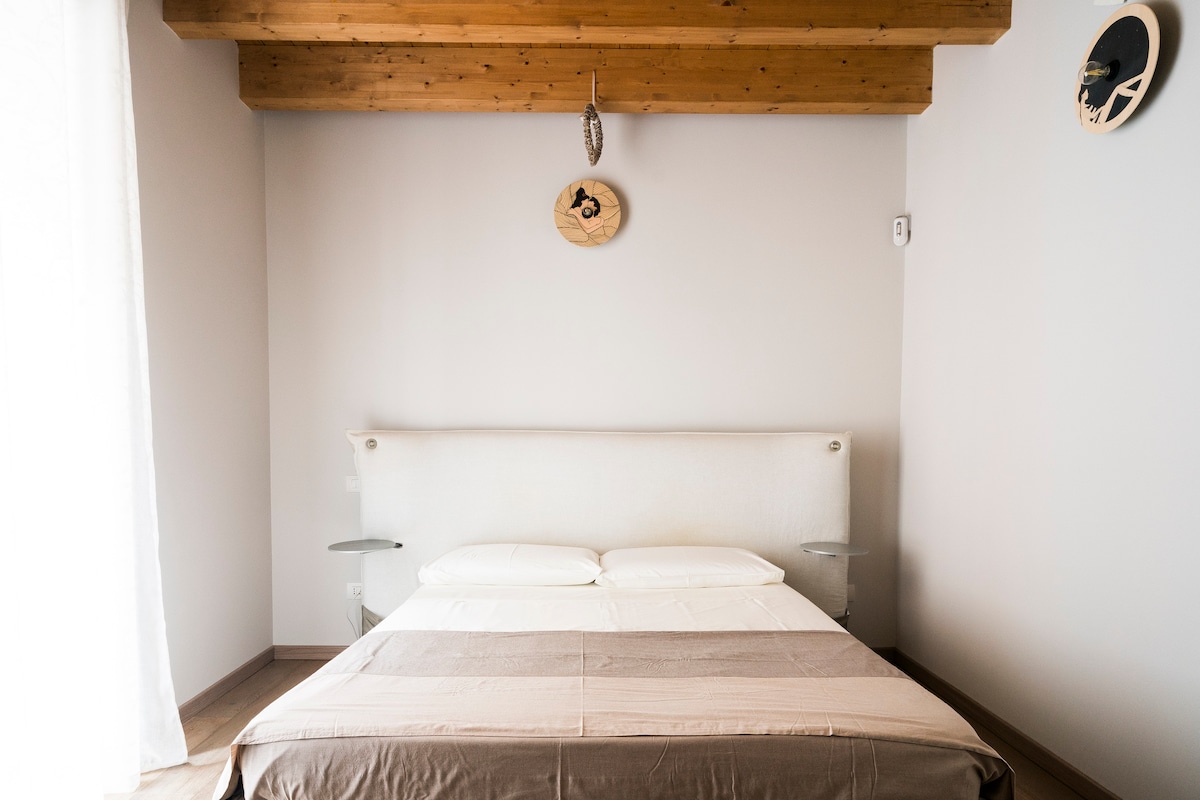 The sleeping area features a spacious bed with a light-colored headboard. Natural light pours in from a nearby window, dressed with sheer curtains. A wooden ceiling adds warmth to the room, complemented by minimalist wall decor.