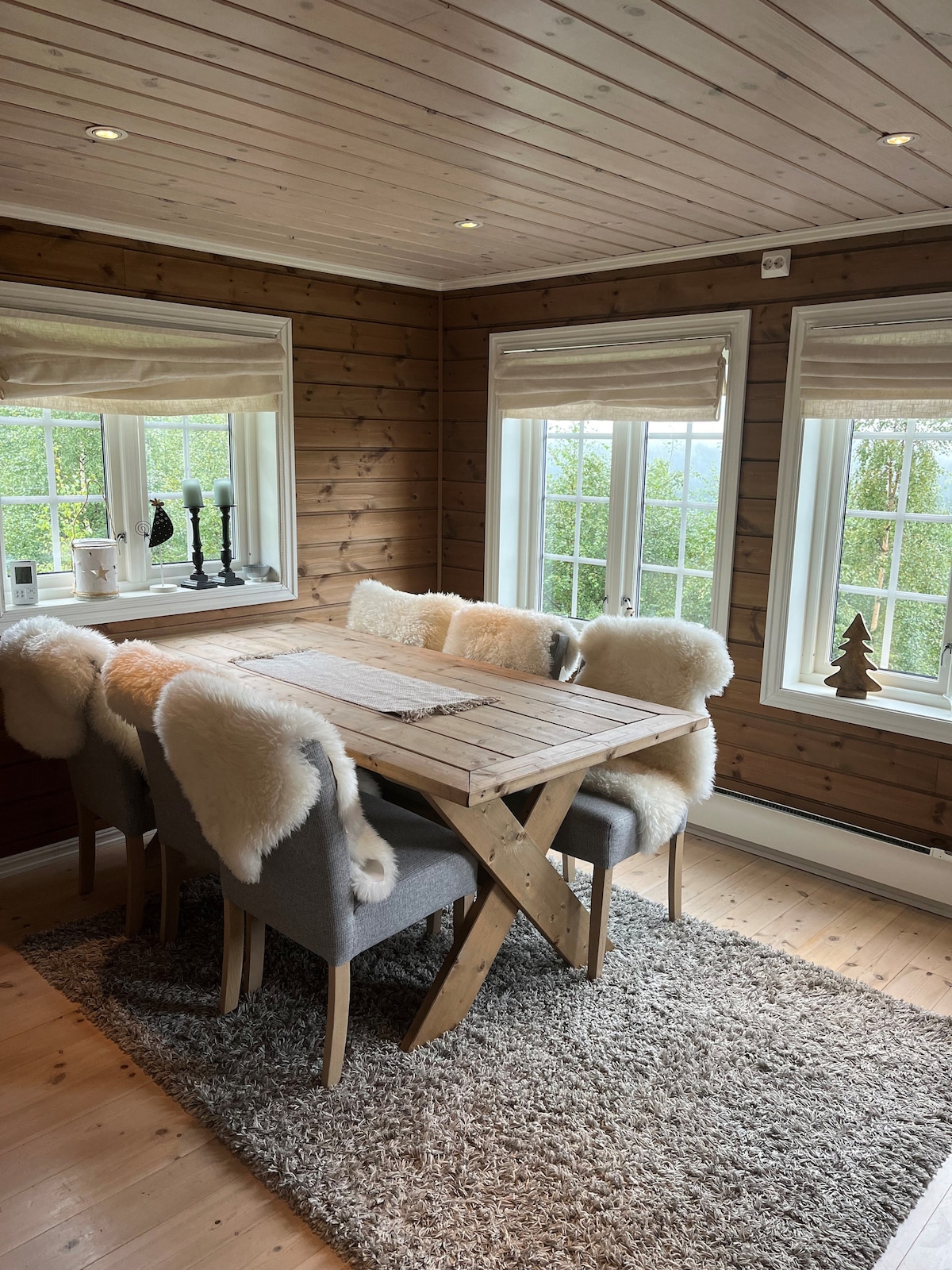 A dining area is featured with a wooden table surrounded by six upholstered chairs. Soft throws cover the chairs, adding comfort. Large windows allow abundant natural light, offering views of the greenery outside. A textured rug lies beneath the table, enhancing the space.