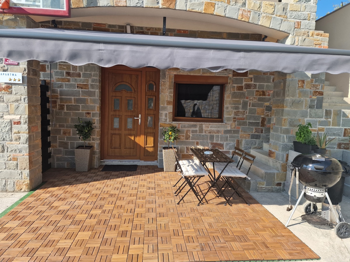 The entrance features a wooden door with decorative glass panels, complemented by stone walls. A small outdoor dining set is positioned on a wooden deck, accompanied by potted plants. A grill is located nearby, offering options for outdoor cooking.