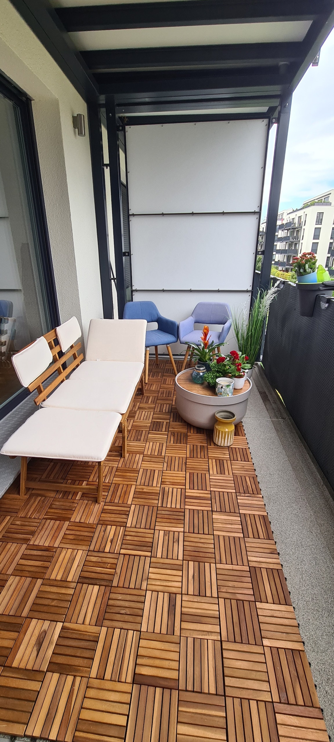 A spacious balcony features wooden flooring and a combination of seating options, including two chairs and a long bench. Potted plants with vibrant flowers are arranged along the railing, complementing the greenery. The view of the courtyard is visible, offering a relaxing outdoor space.