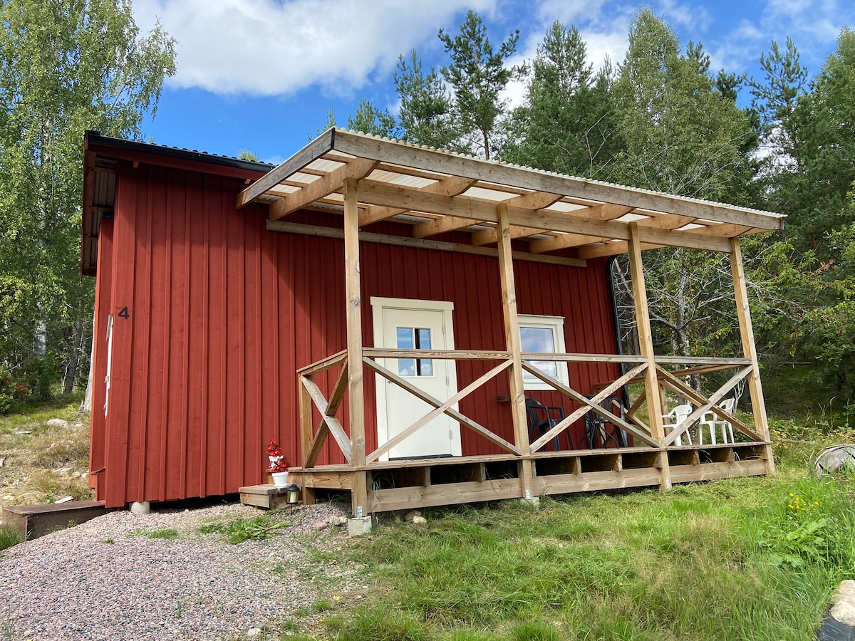 The exterior of a red cabin is presented, featuring a covered porch with a wooden railing. Two white doors provide access to the interior, while several outdoor chairs are positioned on the porch. Surrounding greenery is visible, contributing to the natural setting.