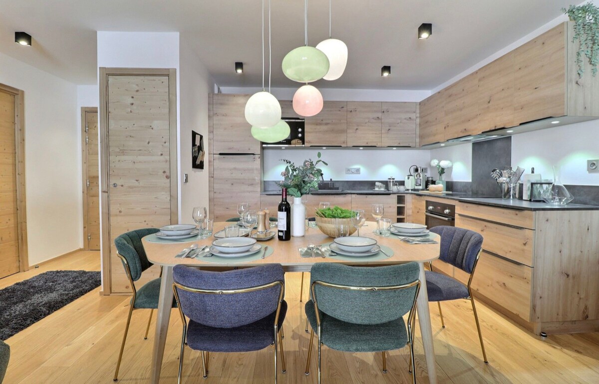 A modern dining area is seen, featuring a wooden table set for eight with elegant dishware and wine. Upholstered chairs in varying colors complement the light wood cabinetry and contemporary pendant lights above. The open kitchen showcases state-of-the-art appliances and provides a welcoming space for dining.