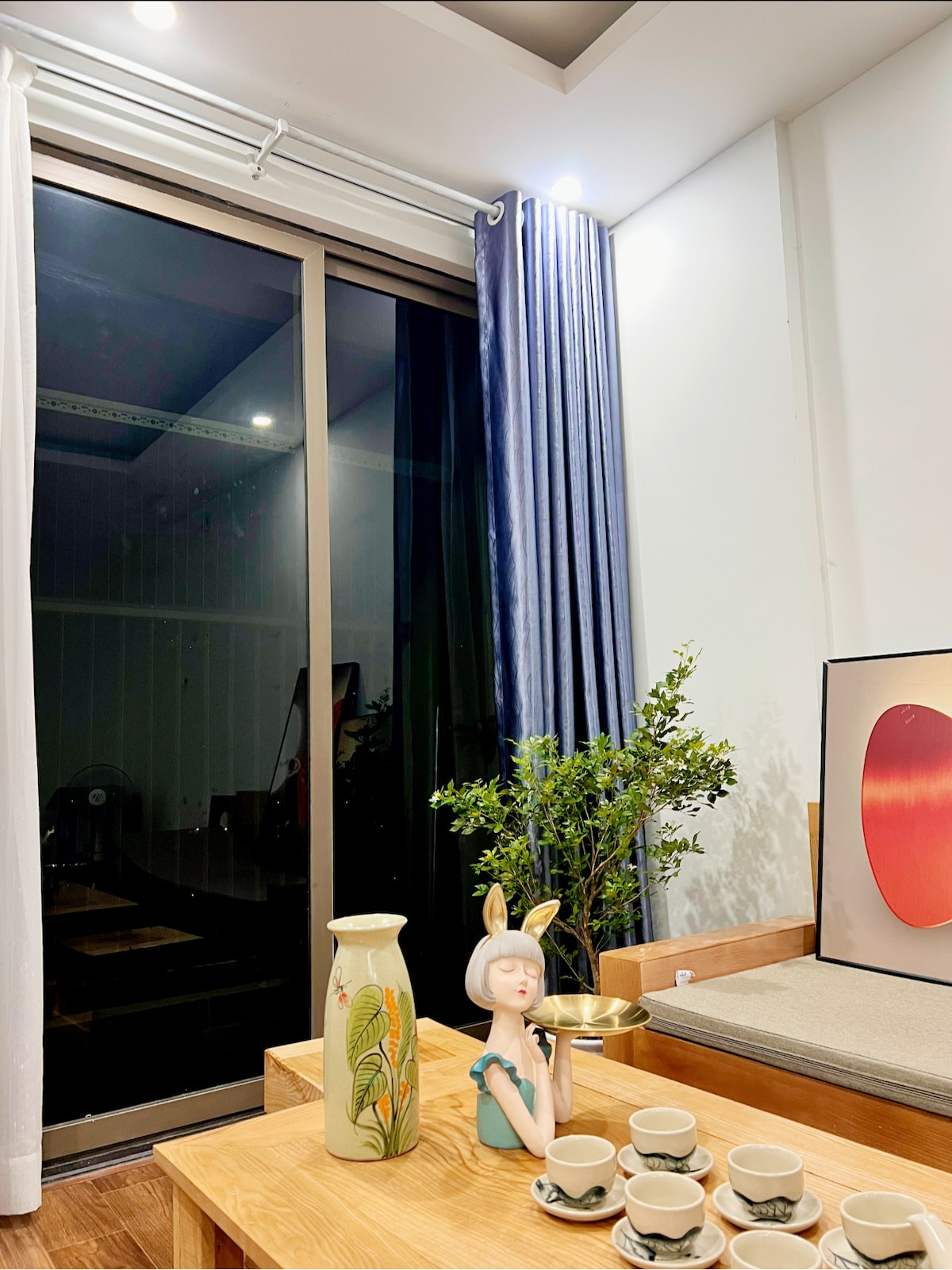 A stylish indoor space features a large glass window draped with soft curtains, allowing natural light to illuminate the area. A wooden coffee table displays a set of decorative cups, a vase with a floral design, and an art piece showcasing warm hues. Greenery adds a calming touch.