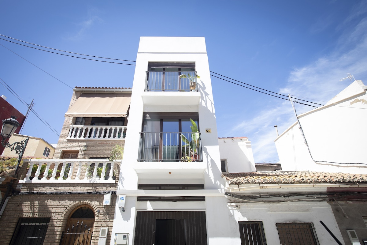 The exterior view of a multi-story house is presented, showcasing a modern white façade. Balconies adorned with plants line the front. Clear blue skies and a few wispy clouds create a vibrant backdrop, while traditional architectural elements contrast with the contemporary design.